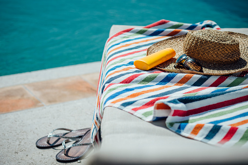Selection of womens items by a pool. There is a colourful towel on the deck chair with a sunhat, sunglasses and sun lotion and some flip flops.