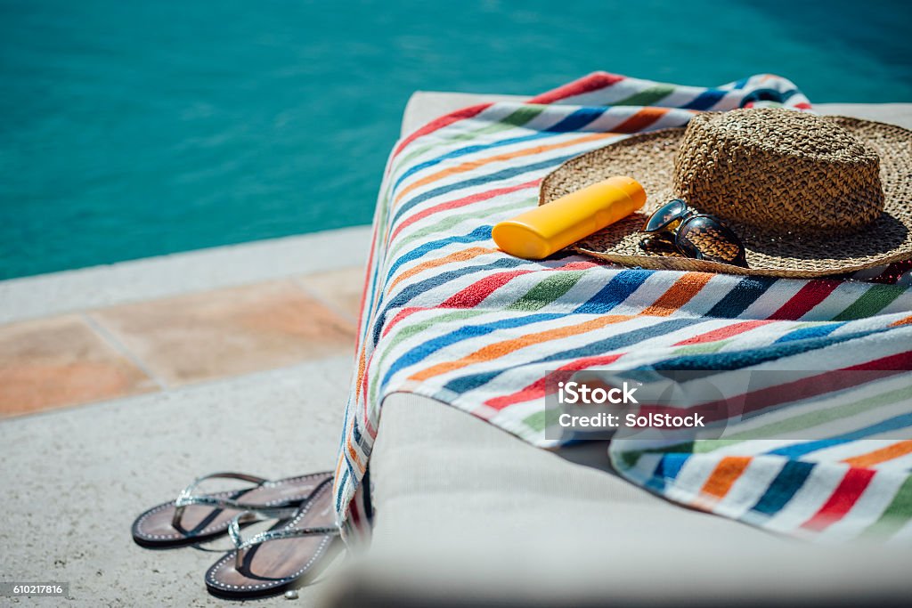 Feminine Pool Necessities Selection of womens items by a pool. There is a colourful towel on the deck chair with a sunhat, sunglasses and sun lotion and some flip flops. Swimming Pool Stock Photo