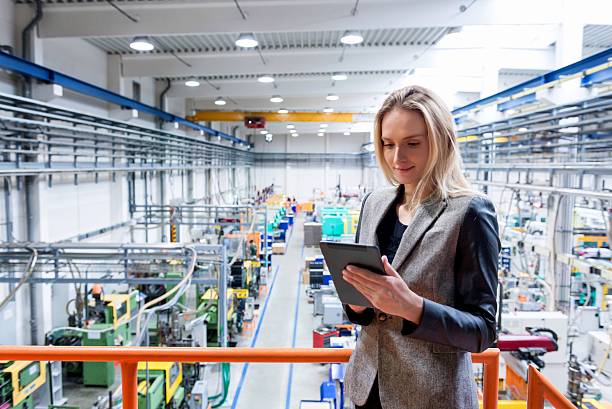 Female inspector in the industrial production! Horizontal color image of blond business female worker standing on balcony on top of large factory, holding digital tablet and examining the production online. Focus on attractive businesswoman, futuristic machines in background. foreman stock pictures, royalty-free photos & images