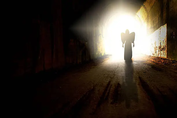 The silhouette of an angel walking toward the light at the end of a tunnel.