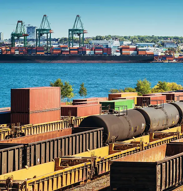 A shunting yard is full of a variety of rail cars in the foreground of a massive container ship in the distance.