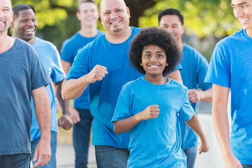 A multi-racial group of boys and men walking or jogging in a park, wearing blue shirts. The focus is on a mixed race African American and Pacific Islander 12 year old boy.