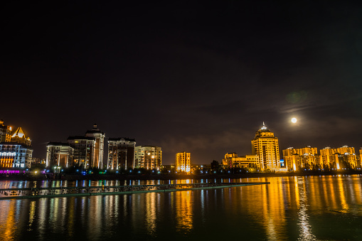 Astana, Kazakhstan. Ishim river embankment in the moon night with buildings and its reflection in the water