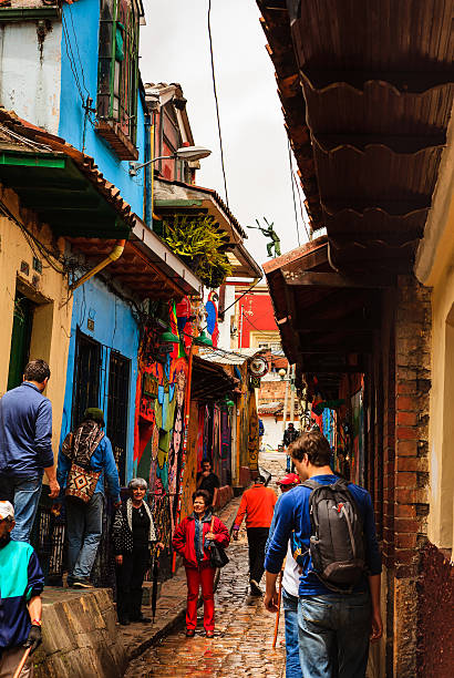 Bogotá, Colombia - Tourists, Both Foreign and Colombian, On The Narrow, Colorful, Cobblestoned Calle del Embudo In The Historic La Candelaria District Bogota, Colombia - July 20, 2016: Tourists on the narrow, cobblestoned, Carrera Segunda that leads to the small square of Chorro de Quevedo, in the historic district of La Candelaria. A local tour guide, the man on the left, with the Arhuaca Mochila - a bag originally made by the Arhuaca tribe of the Sierra Nevada, is with the group. The capital city of Colombia, Bogota, was founded in the 16th Century in this area, by the Spanish Conquistador, Gonzalo Jiménez de Quesada. Many walls in this area are painted with either street art, or legends of the pre Colombian era, in the vibrant colours of Latin America. The sky is overcast. Photo shot in the morning sunlight, on a cloudy day. Vertical format. calle del embudo stock pictures, royalty-free photos & images