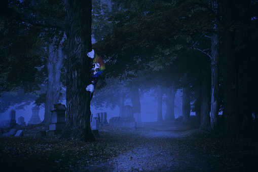 A creepy clown hiding behind a tree in a cemetery at night.