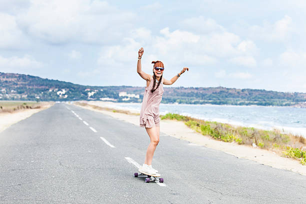 Cheerful young woman skateboarding on road next to the sea Cheerful young boho woman skateboarding on road next to the sea. Laughing, hands up. With casual clothes, headband, sunglasses and hair in braids, bracelets. Landscape view on background. longboarding stock pictures, royalty-free photos & images
