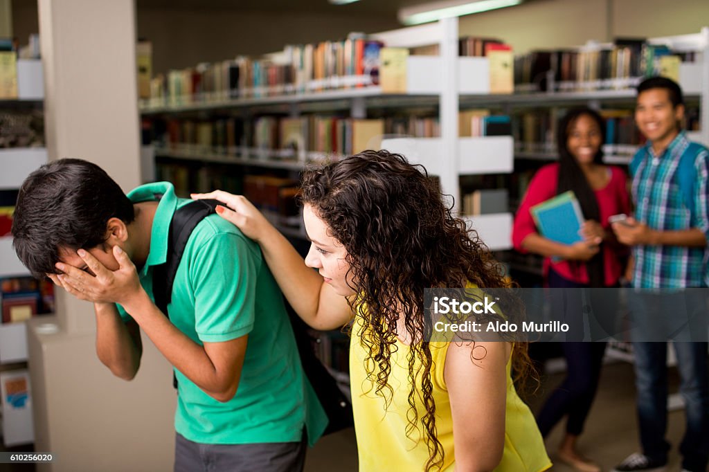 Female student helping sad friend A latin female student helping his sad friend at the library in a horizontal medium shot indoors with people in the background. Bullying Stock Photo