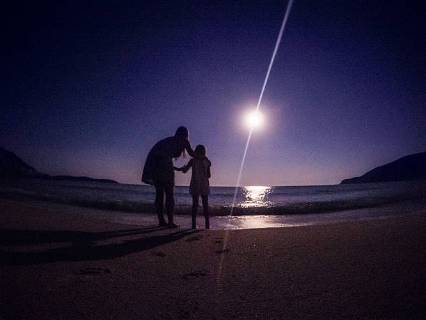 Mother is holding her child on the beach at night while both are enjoying the full moon, reflecting on the sea. Entirely made by the moonlight. Features prominent film grain.