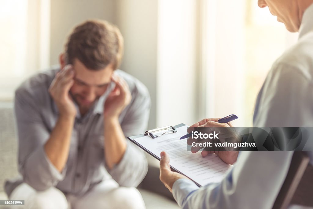 Man at the psychotherapist Handsome depressed man in casual clothes is leaning on his knees and telling about his problems while sitting on the couch at the psychotherapist. Doctor in focus Mental Health Stock Photo