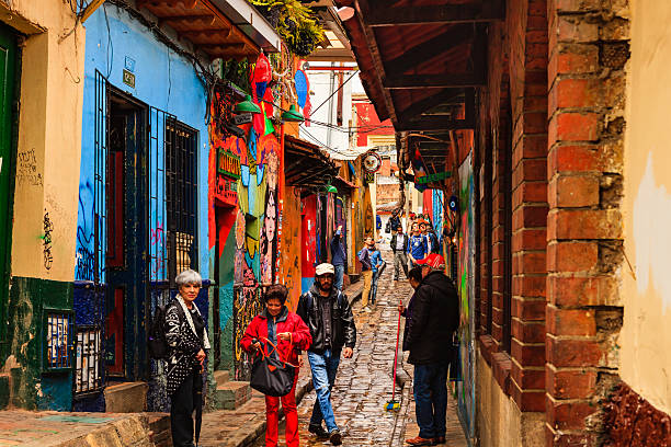 Bogotá, Colombia - Local Colombian Tourists Walk Through The Narrow, Colorful, Cobblestoned Calle del Embudo In The Historic La Candelaria District Bogota, Colombia - July 20, 2016: Tourists both local and international, on the narrow, cobblestoned, Carrera Segunda that leads to the small square of Chorro de Quevedo, in the historic district of La Candelaria. The Andean capital city of Colombia, Bogota, was founded in the 16th Century in this area, by the Spanish Conquistador, Gonzalo Jiménez de Quesada. Many walls in this area are painted with either street art, or legends of the pre Colombian era, in the vibrant colours of Latin America. The sky is overcast. It has just rained a little and the Carrera is wet. Photo shot in the morning sunlight, on a cloudy day. Horizontal format. calle del embudo stock pictures, royalty-free photos & images