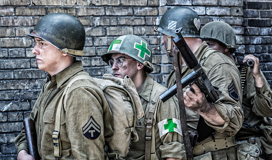 A heavily armed platoon of World War II US Army soldiers is standing in line against an ancient brick wall as they wait solemnly and anxiously before moving out on a patrol mission. The technical specialist in front is carrying an M1 Garand .30 caliber rifle. He is followed by the team's combat medic with the medical cross symbols (changed to green to avoid infringing American Red Cross trademark rights) on his helment and armband; then the unit patrol leader Sergeant - holding an M1928Ai .45 caliber Thompson submachine gun; finally the last soldier carrying an M1919 Browning machine gun with a bandoleer of .30 caliber ammunition.