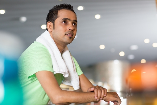 Close up of young Indian man with towel around neck leaning on glass fence while looking somewhere tiredly with blurred view of fitness center hall in background