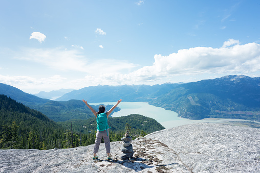 A young, female Asian backpacker stands next to a stone cairn on a mountaintop in Squamish, British Columbia, Canada.  View of the valley and surrounding mountains in the background.