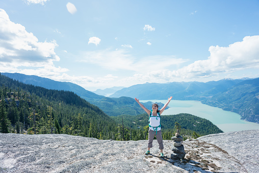 A young, female Asian backpacker with arms raised stands next to a stone cairn on a mountaintop in Squamish, British Columbia, Canada.  View of the valley and surrounding mountains in the background.
