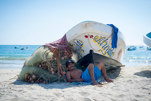 Puerto Escondido, Mexico - November 5, 2014: A Mexican fisherman relaxes in the shade of a boat on the beach in Puerto Escondido, Mexico.