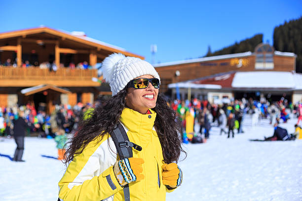Cheerful young woman standing in front of a ski chalet. Wears ski wear, ski goggles, gloves and knit hat, with backpack. On background tourists in front of a ski chalet. Focus on foreground.