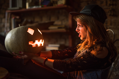 Angry woman holding Jack O' Lantern in her hands and looking at it while having a furious facial expression.