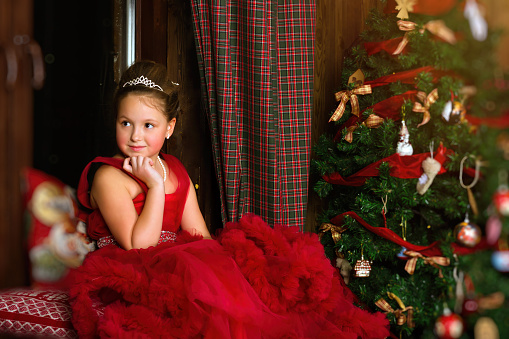 Little winter Princess - girl in red dress, sitting on windowsill, looks out window at night. Celebration of New year and Christmas in enchanting holiday interior with decorated pine