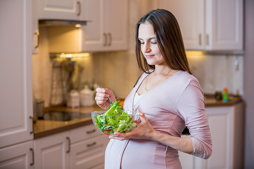 A young beautiful pregnant woman eating a fresh green salad. Healthy nutrition and pregnancy. Pregnant woman's standing in the kitchen with vegetable salad. Healthy lifestyle.