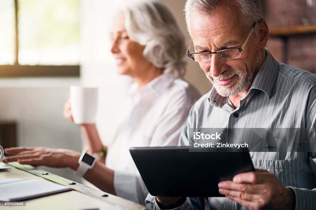 Elderly couple together at the kitchen Daily life. Old grey man in glasses using laptop while woman drinking tea and smiling Senior Adult Stock Photo