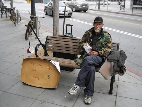 Los Angeles, CA, United States - 16 June 2010. Unemployed man sitting on a bench and begging for money. Very witty writing on the carton reads: he catches fish - money.