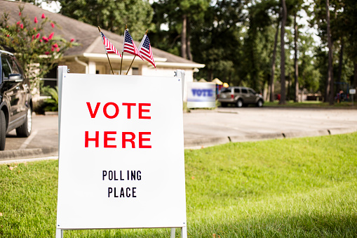 'Vote Here, Polling Place' sign outside of a local polling location in USA.  American flags top the sign.  Parking lot and a public community center building in background. The USA elections are held in November each year.  No people.