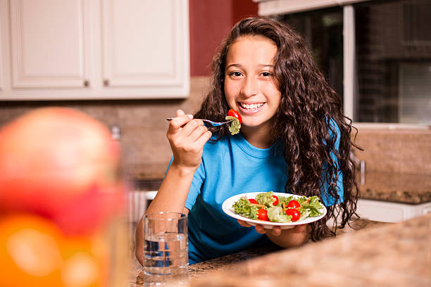 Teenage girl eating salad for dinner after school. Day in the life of a teenager. A Latin descent teenage girl eats healthy salad after her school soccer team practice. She has a plate full of lettuce, carrots and tomatoes and also has a glass of water. This image is part of a series where our model wakes up, eats breakfast, heads to school, comes home, plays soccer, eats dinner, completes her homework, and goes to bed. Teenager EatING stock pictures, royalty-free photos & images