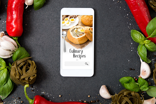 Smartphone with culinary recipes application lying on stone counter and surrounded by fresh herbs and hot spices