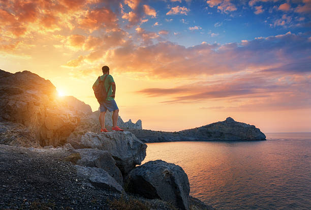 Beautiful summer landscape with standing man with backpack on the stone at the ocean against the colorful sky with clouds at sunset. Travel background. Sport, lifestyle. Tourism