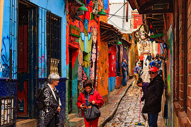 Bogotá, Colombia - Tourists On the Narrow, Colorful, Cobblestoned Calle Del Embudo In The Historic La Candelaria District Of The Capital City Bogota, Colombia - July 20, 2016: Tourists both local and international, on the narrow, cobblestoned, Carrera Segunda that leads to the small square of Chorro de Quevedo, in the historic district of La Candelaria. The Andean capital city of Colombia, Bogota, was founded in the 16th Century in this area, by the Spanish Conquistador, Gonzalo Jiménez de Quesada. Many walls in this area are painted with either street art, or legends of the pre Colombian era, in the vibrant colours of Latin America. The sky is overcast. It has rained a little and the Carrera is wet. Photo shot in the morning sunlight, on a cloudy day. Horizontal format. calle del embudo stock pictures, royalty-free photos & images
