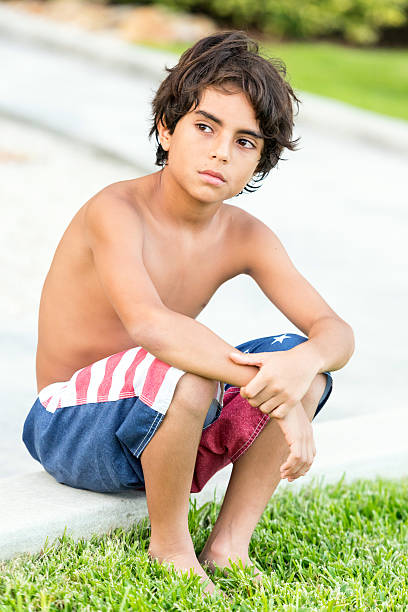 Hispanic eleven years old  little boy wearing a swim suit sitting on the floor looking away