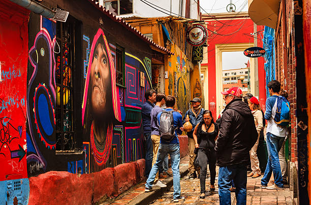 Bogotá, Colombia - Tourists, Both Foreign and Colombian, Walk Through The Narrow, Colorful, Cobblestoned Calle del Embudo In The Historic La Candelaria District Bogota, Colombia - July 20, 2016: Tourists both local and international, on the narrow, cobblestoned, Carrera Segunda, almost at the point where it leads into the small square of Chorro de Quevedo, in the historic district of La Candelaria. The Andean capital city of Colombia, Bogota, was founded in the 16th Century in this area, by the Spanish Conquistador, Gonzalo Jiménez de Quesada. Many walls in this area are painted with either street art, or legends of the pre Colombian era, in the vibrant colours of Latin America. There is a brightly painted shop front on the left of the image. The sky is overcast. Photo shot in the morning sunlight, on a cloudy day. Horizontal format. calle del embudo stock pictures, royalty-free photos & images