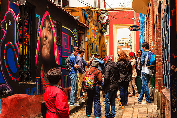 Bogotá, Colombia - Tourists, Both Foreign and Colombian, On The Narrow, Colorful, Cobblestoned Calle del Embudo In The Historic La Candelaria District Bogota, Colombia - July 20, 2016: Tourists both local and international, on the narrow, cobblestoned, Carrera Segunda, almost at the point where it leads into the small square of Chorro de Quevedo, in the historic district of La Candelaria. The Andean capital city of Bogota, Colombia, was founded in the 16th Century in this area, by the Spanish Conquistador, Gonzalo Jiménez de Quesada. Many walls in this area are painted with either street art, or legends of the pre Colombian era, in the vibrant colours of Latin America. There is a brightly painted shop front on the left of the image. The sky is overcast. Photo shot in the morning sunlight, on a cloudy day. Horizontal format. calle del embudo stock pictures, royalty-free photos & images