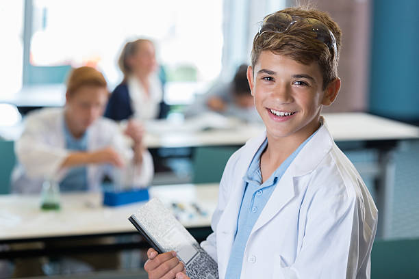 Smiling Hispanic preteen wearing lab coat in science class Preteen Hispanic male student wearing a lab coat and standing in front of a science class while holding a composition notebook. Smiling Hispanic preteen male in science class wearing a lab coat with notebook. high school student child little boys junior high stock pictures, royalty-free photos & images