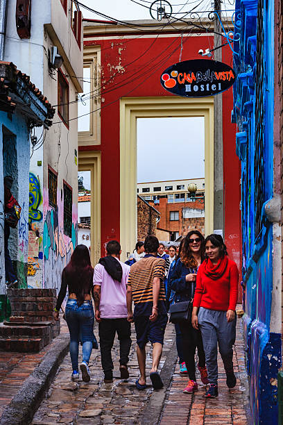 Bogotá, Colombia - The Gateway That Leads To The Chorro De Quevedo At The Narrowest End Of The Colorful, Cobblestoned Calle del Embudo, In The Historic La Candelaria Bogota, Colombia - July 20, 2016: Some local Colombian youth on the narrow, cobblestoned, Carrera Segunda, almost at the point where it leads into the small square of Chorro de Quevedo, in the historic district of La Candelaria. The gateway in the background leads to the square. The Andean capital city of Bogota, Colombia, was founded in the 16th Century in this area, by the Spanish Conquistador, Gonzalo Jiménez de Quesada. Many walls in this area are painted with either street art, or legends of the pre Colombian era, in the vibrant colours of Latin America.  The sky, visible through the gateway, is overcast. Photo shot in the morning sunlight, on a cloudy day. Horizontal format. calle del embudo stock pictures, royalty-free photos & images