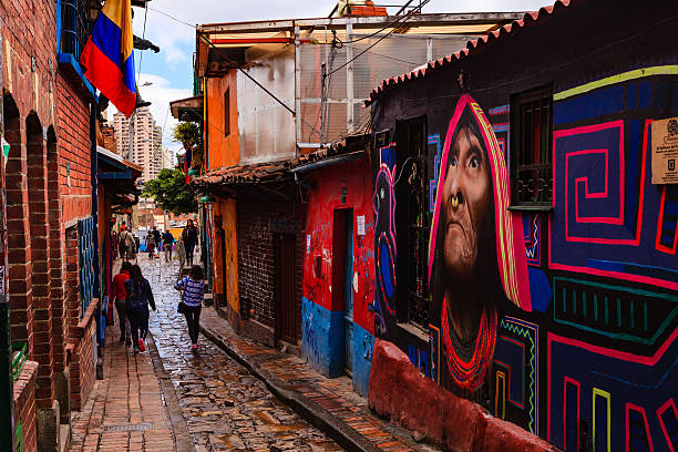 Bogotá, Colombia - People Walk Through The Narrow, Colorful, Cobblestoned Calle del Embudo In The Historic La Candelaria District Bogota, Colombia - July 20, 2016: Some local Colombian people walk down the narrow, cobblestoned, Carrera Segunda, in the historic district of La Candelaria, in the Andean capital city of Bogota, Colombia, in South America. The City was founded in the 16th Century in this area, by the Spanish Conquistador, Gonzalo Jiménez de Quesada. Many walls in this area are painted with either street art, or legends of the pre Colombian era, in the vibrant colours of Latin America.  The wall of a shop on the right is an example of such murals. It has rained a little and the Carrera is wet; the overcast sky promises more rain. 20th July is National Day in Colombia and the country's flag is flown on many buildings. Photo shot in the morning sunlight, on a cloudy day. Horizontal format. calle del embudo stock pictures, royalty-free photos & images