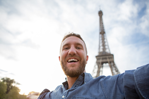 Young cheerful man near the Eiffel tower in Paris-France take a selfie portrait using a mobile phone. Beautiful summer day.