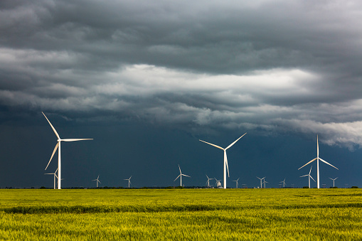 Dark black storm clouds pass over a row of clean renewable energy wind turbines in the middle of a grain crop.  In the background, distant grain storage.  Horizontal, copy space, metaphor.