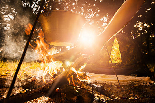 Unrecognizable man preparing food on camping at sunset and adding wood logs on campfire.
