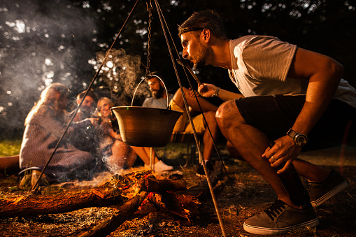 Side view of man cooking and tasting food from cauldron while camping in the forest with his friends.