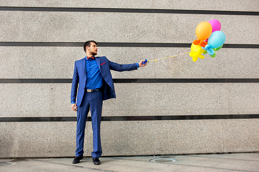 Young businessman standing against the wall with bunch of multi colored balloons. Copy space.