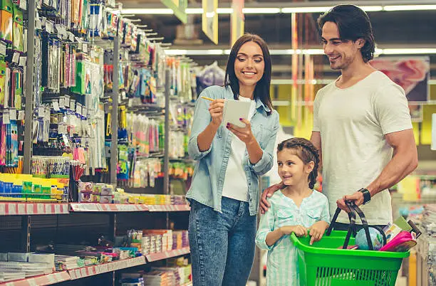 Beautiful young parents and their cute little daughter are smiling while choosing school stationery in the supermarket. Mom is making notes in the list