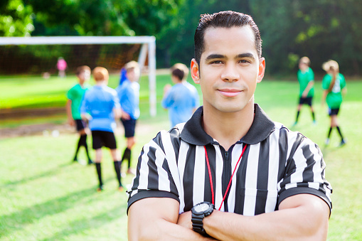 Handsome mid adult Hispanic referee stands on the field during soccer game. He has his arms crossed and is wearing referee uniform. The soccer teams are in the background along with soccer goal.
