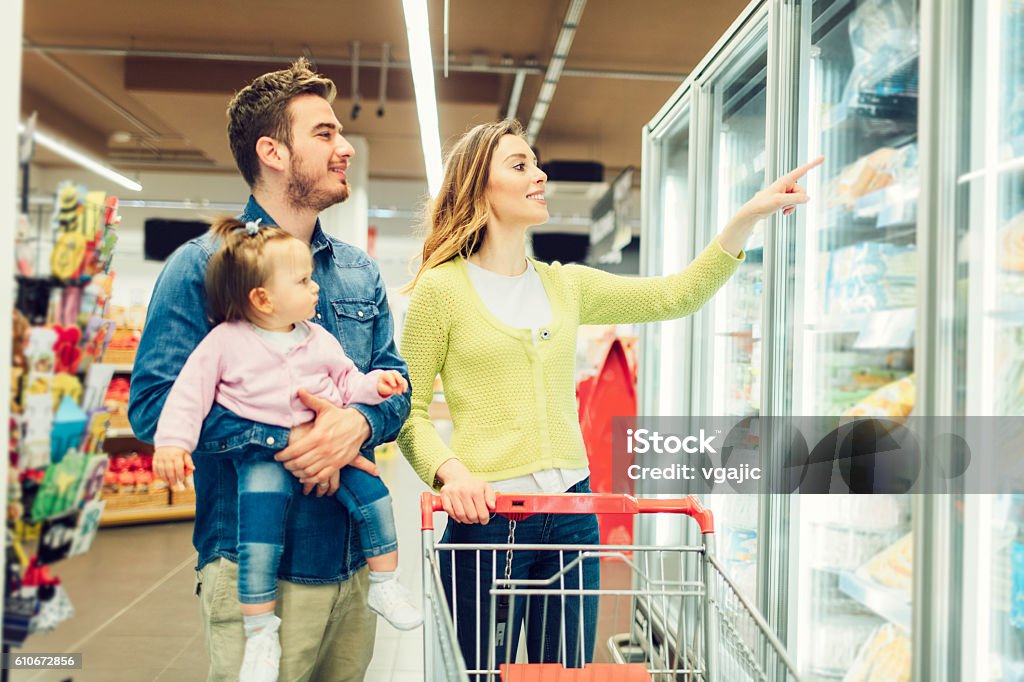 Mother And Father With Their Baby Daughter Grocery Shopping Mother and father and their baby girl in groceries shopping. Father carry his little baby daughter and they are choosing frozen food. They are happy together.  Location released. Frozen Food Stock Photo