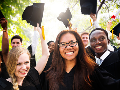 A group of multi-ethnic students graduating from high school or college, standing in  row outdoors, wearing black caps and gowns. The focus is on the young black woman smiling confidently at the camera.