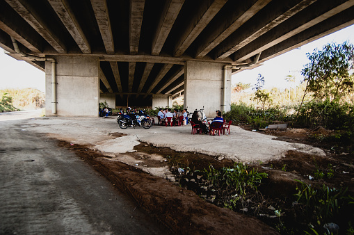 Ho Chi Minh City, Vietnam - February 5, 2015: View from under Phu My bridge, one of the biggest bridges in Ho Chi Minh City. Days get very hot and the whole car and city concrete just makes it more hotter, so locals gather to relax in the eternal shade under Phu My bridge, it is pretty cool and humid under there because this place is near Saigon river. Sitting on their small  chairs, scooters parked near by and eating local Pho soup.
