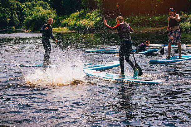 She fell in! Group of teenagers having fun during a paddleboarding class. They are all laughing at one person who has fallen in, making a big splash. immersed in their work stock pictures, royalty-free photos & images