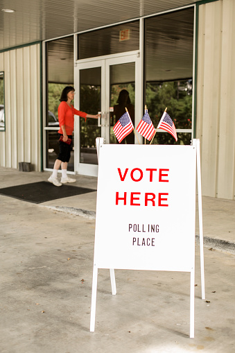'Vote Here, Polling Place' sign outside of a local, public polling location in USA.  American flags top the sign.  One woman enters the building.  The USA elections are held in November each year.