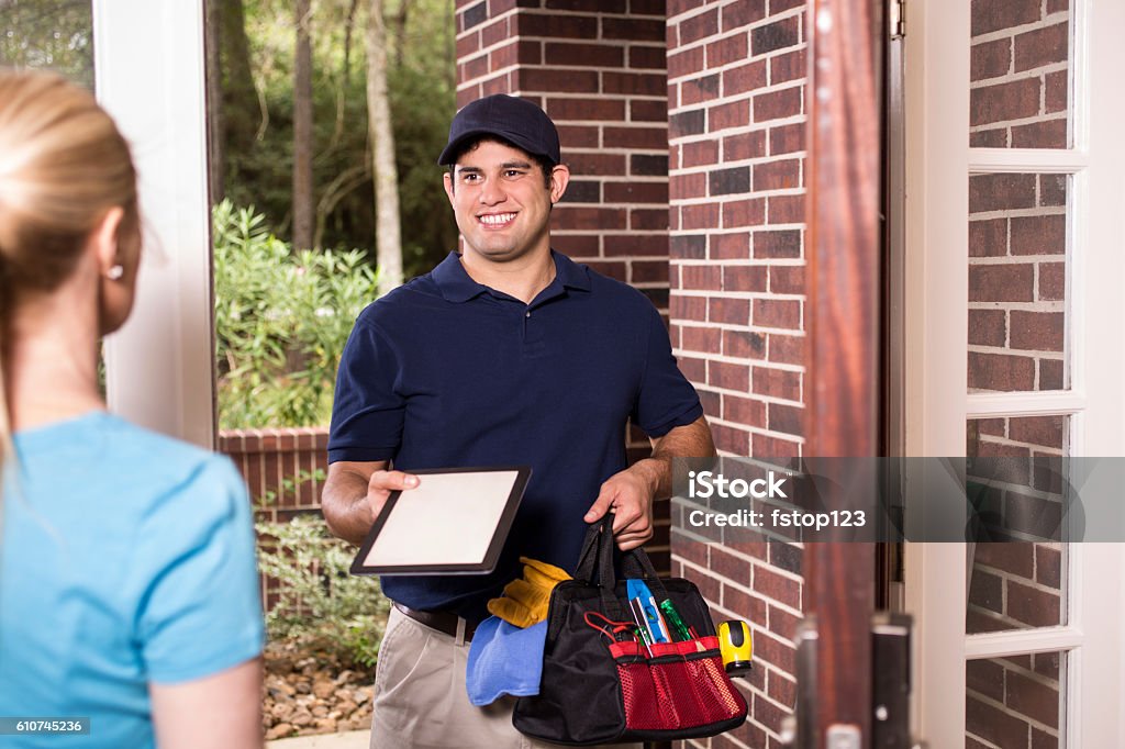 Repairman at customer's front door. Latin descent repairman or blue collar/service industry worker makes service/house call at customer's front door. Man holds digital tablet and toolbag.  Inspectors, exterminators, plumbers, electricians.  Red brick home. Plumber Stock Photo