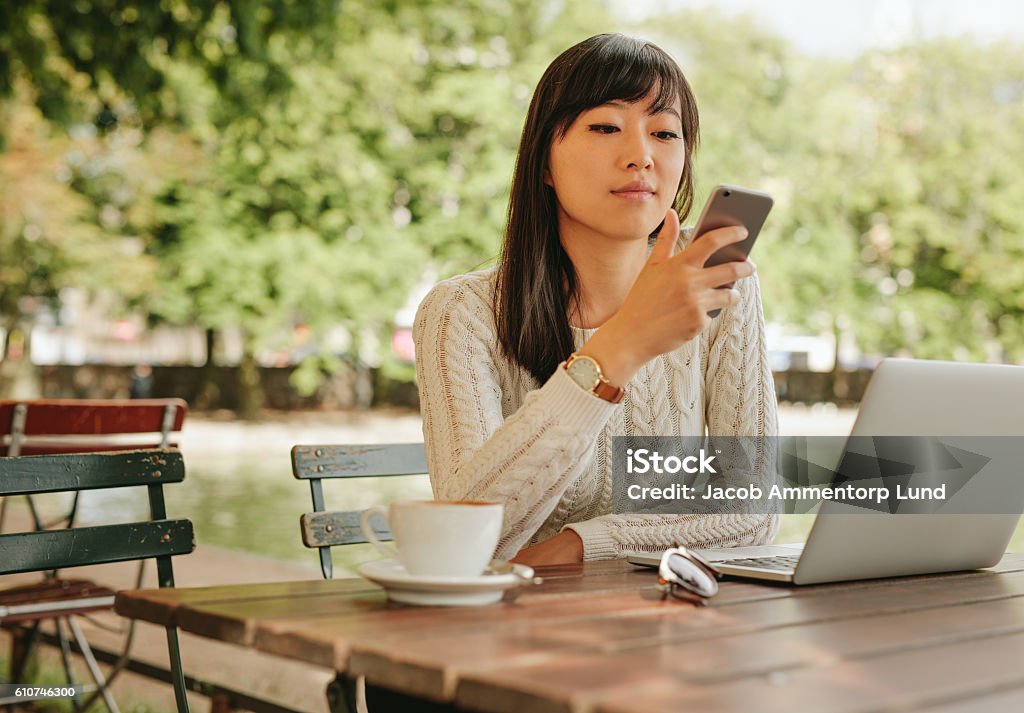 Attractive woman using smartphone at coffee shop Shot of attractive asian woman using smartphone at outdoor coffee shop. Female sitting at cafe table outdoors using mobile phone. Cafe Stock Photo